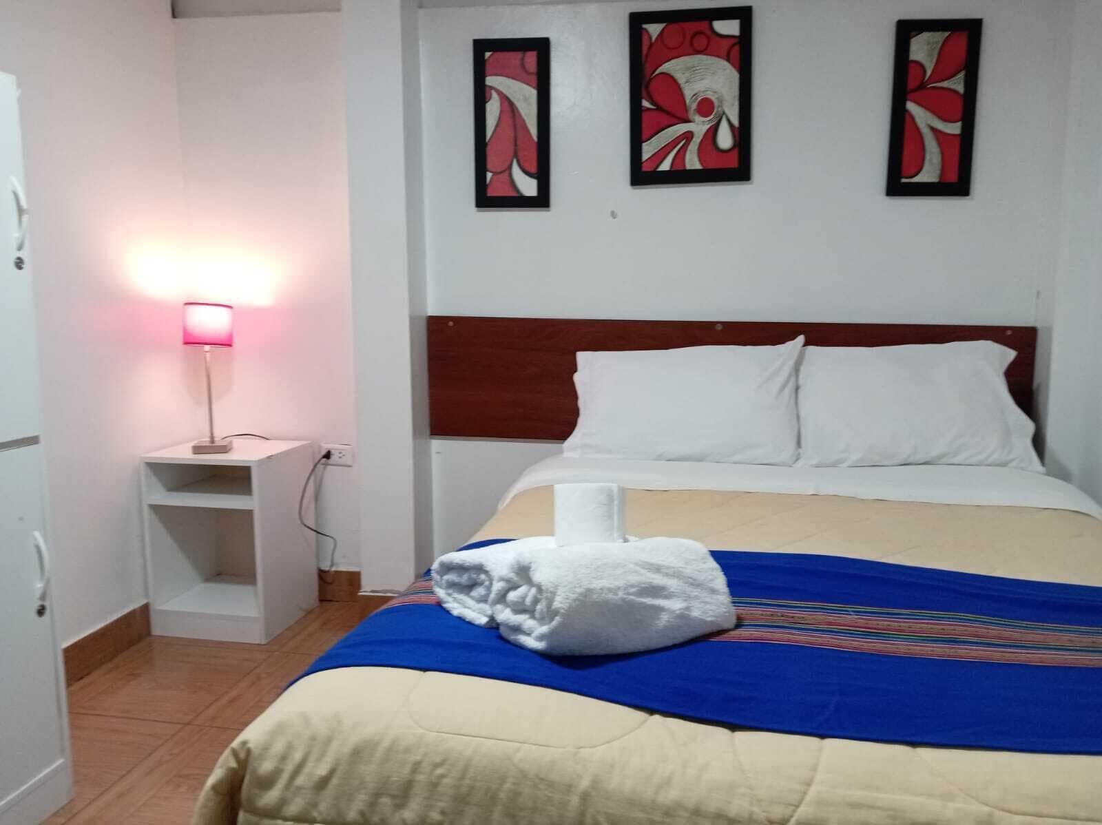 A hotel room with a bed, a nightstand with a lamp, and three abstract wall art pieces.