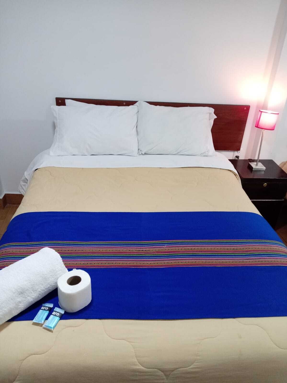 A bed with white pillows, a blue and beige bedspread, a nightstand with a lamp, and toiletries.