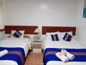 Ein Doppelzimmer mit zwei Betten, jeweils mit einem hölzernen Kopfteil, weißer Bettwäsche und blau gestreiften Decken. Zwischen den Betten steht ein Nachttisch mit einer Lampe, und ein kleiner Tisch mit einem dekorativen Gegenstand.