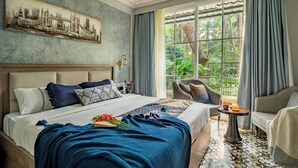 Habitación de hotel con una cama grande, un cuadro de un paisaje urbano, vistas a zonas verdes por la ventana y una mesita con una bebida.
