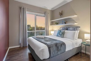 Ein Schlafzimmer mit einem großen Bett, einem Fenster mit Vorhängen, einem Nachttisch mit Lampe und einem Regal mit Büchern und einer Pflanze.