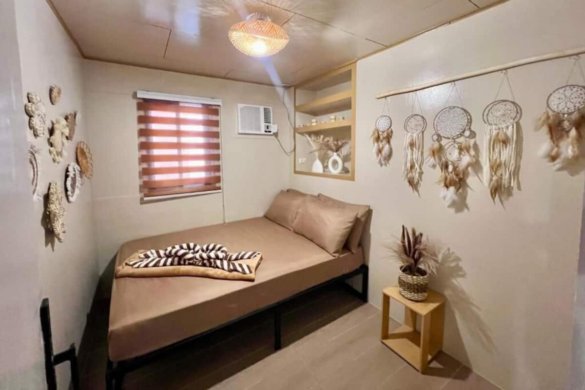 Un dormitorio con una cama, una ventana con persianas, un aire acondicionado de pared, una mesita de noche con una planta y atrapasueños en la pared.