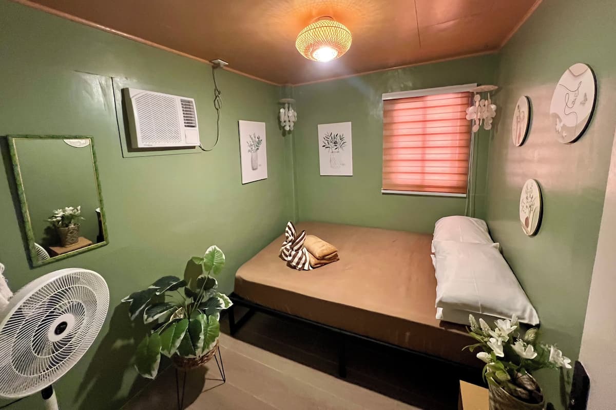 Una habitación con cama, ventilador de pie, planta en maceta y adornos en la pared.