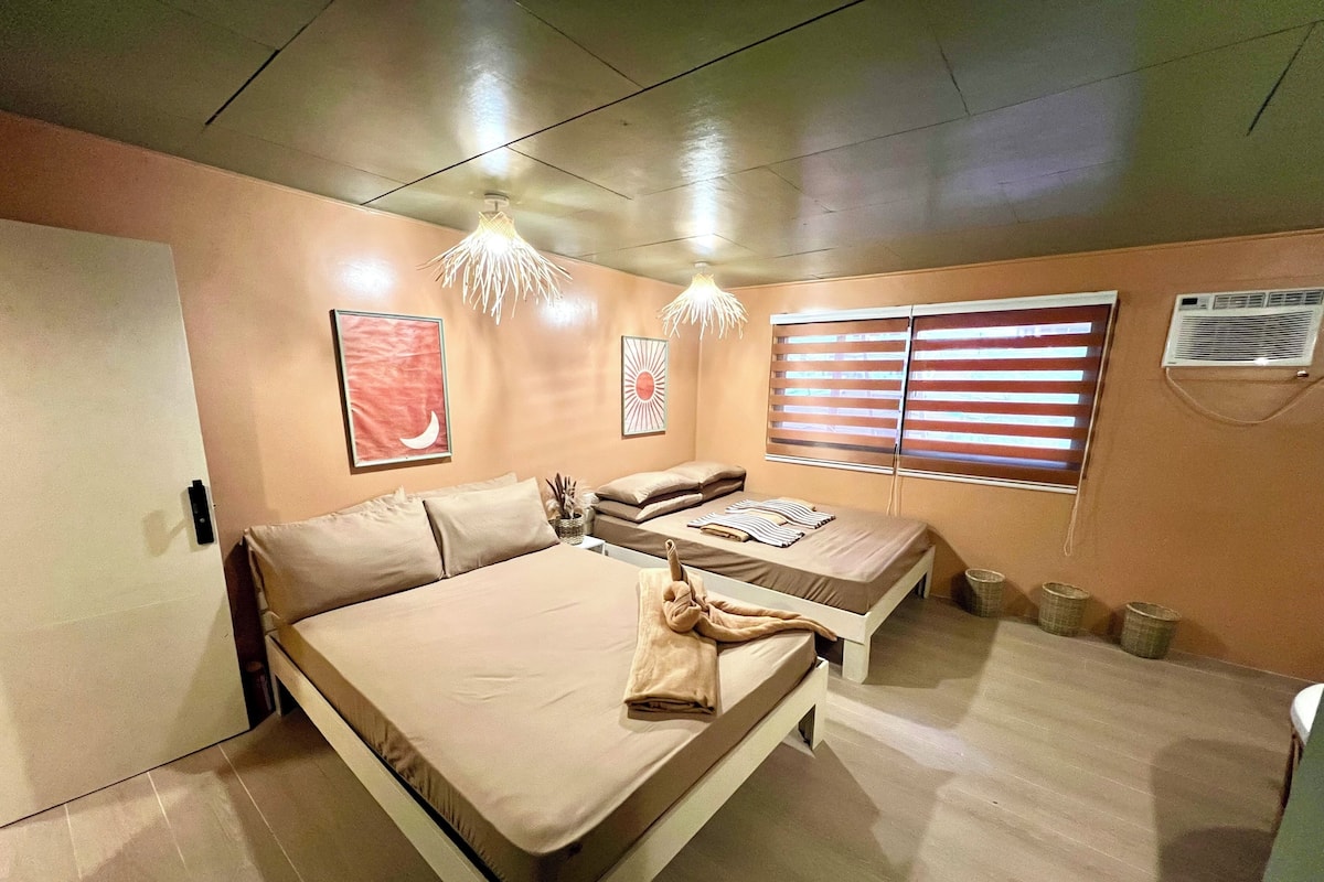 Un dormitorio con cama, dos mesitas de noche, una ventana con persianas, un aire acondicionado de pared y una lámpara en el techo.