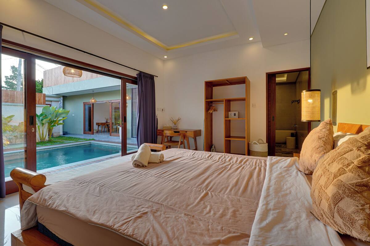 Ein Schlafzimmer mit einem großen Bett, einem kleinen Schreibtisch, einem Stuhl, einem Badezimmer und Blick auf einen Pool.