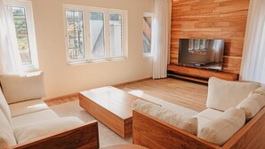 A modern living room with a sofa, coffee table, and TV. There are windows with curtains, a wooden wall, and a view of a building. - Stone Heaven Villa (Nuwara Eliya)