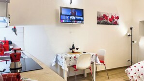 Een kleine kamer met een tv aan de muur, een tafel met stoelen, een bed en een kitchenette met een spoelbak en keukengerei.