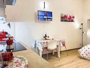 Ein kleines Zimmer mit Fernseher an der Wand, einem Tisch mit Stühlen, einem Bett und einer Küchenzeile mit Spüle und Geschirr.