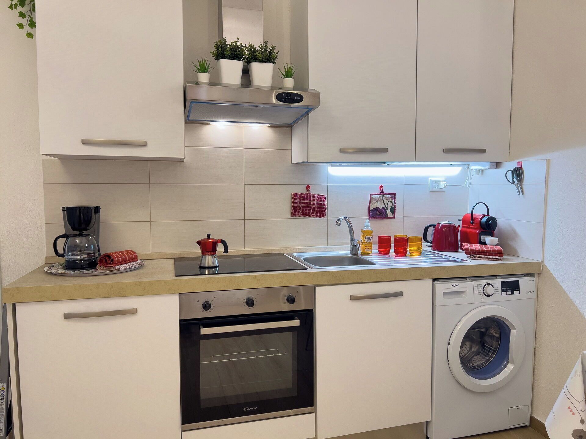 A compact kitchen with a coffee maker, kettle, sink, and a washing machine.