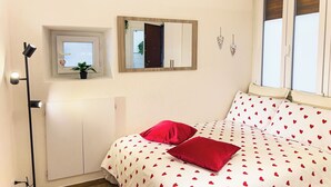 Een slaapkamer met een bed, een bedlampje, een klok, een spiegel en een raam met jaloezieën.