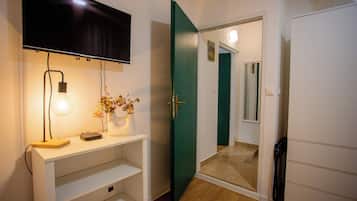A room with a TV mounted on the wall, a desk with a lamp, a vase with flowers, and a green door leading to another room.
