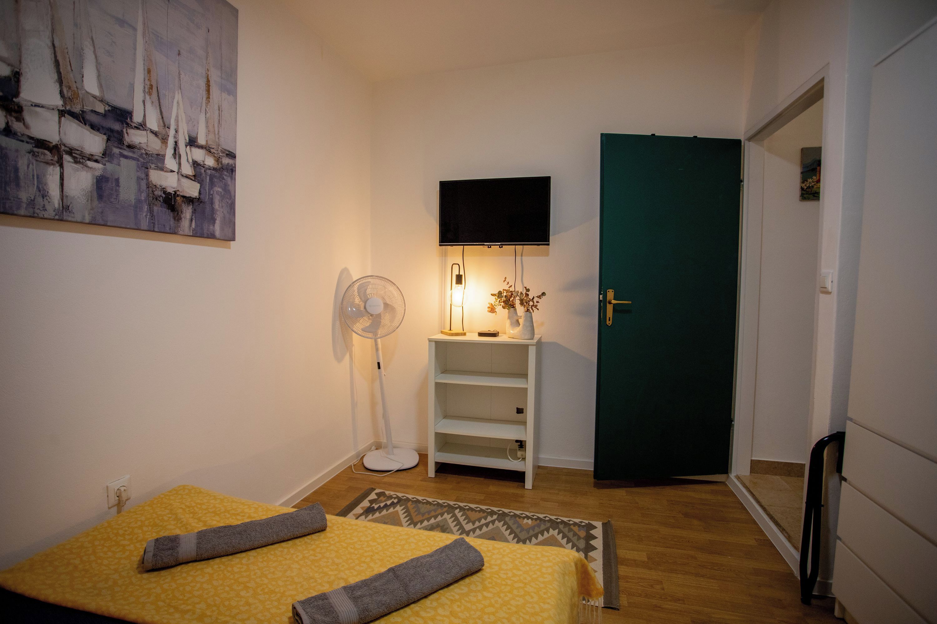 A hotel room with a bed, a standing fan, a TV mounted on the wall, and a door.