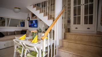 A dining area with a white table set for two, a staircase with a wooden handrail, and a television mounted on the wall.
