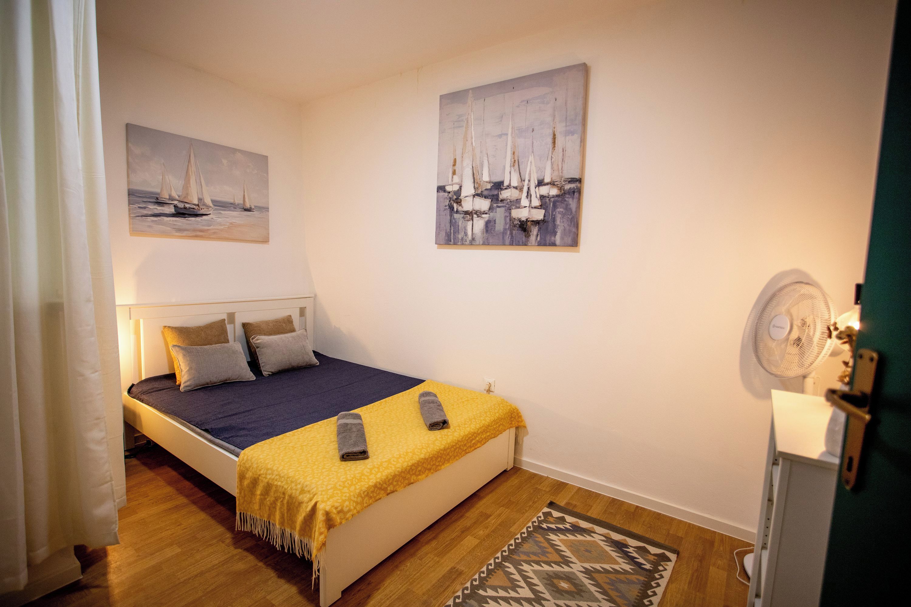 A single bed with a blue and yellow bedspread, two pillows, a small rug, and two remote controls on the bed.