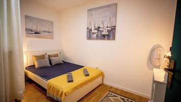 A single bed with a blue and yellow bedspread, two pillows, a small rug, and two remote controls on the bed.