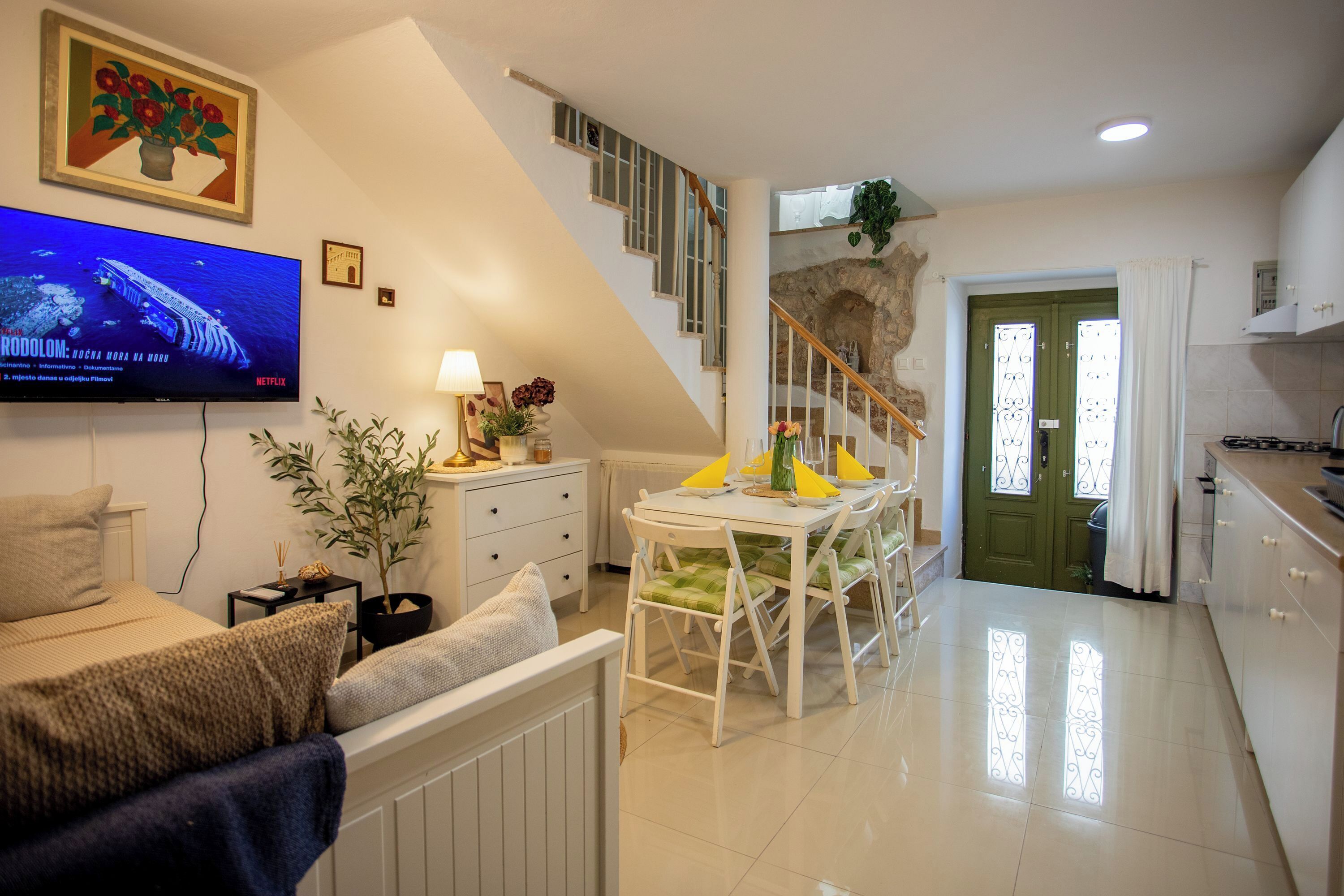 A modern living room with a flat-screen TV, a dining area with a table and chairs, a staircase, and a green door.