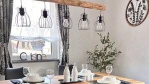 Un espace repas avec une table en bois, des chaises noires, des lampes suspendues et une vue sur l’extérieur.