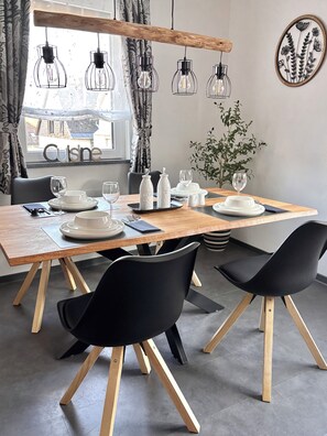 A dining area with a wooden table, black chairs, hanging pendant lights, and a view of the outside. - Ferienhaus Schieferland - Your vacation home between the Moselle and the Nürburgring (Müllenbach)