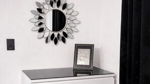 Une commode blanche, surmontée d’un miroir et d’un cadre photo, est placée contre un mur blanc, devant un rideau noir.