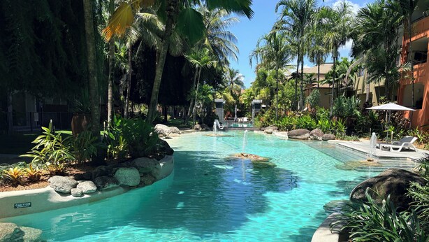 Uma piscina cristalina rodeada por pedras e plantas tropicais, com espreguiçadeiras e guarda-sóis.