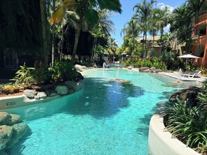 A clear pool surrounded by rocks and tropical plants, with lounge chairs and umbrellas. - Resort Style Apartment Close to the City with 9 pools! (Westcourt)