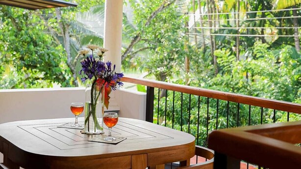 Una mesa de madera redonda con dos vasos de líquido ámbar, un florero con flores púrpuras y vistas a follaje exuberante.