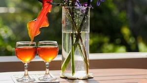 Dos vasos de líquido ámbar sobre una mesa con un florero transparente que contiene flores moradas y una hoja roja.
