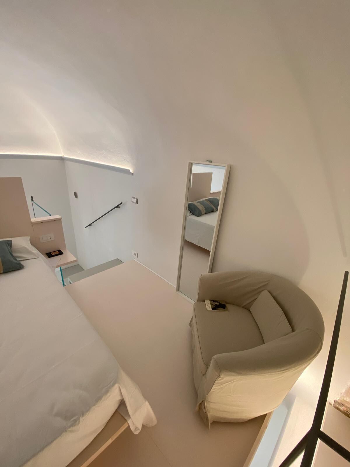 Un dormitorio moderno con cama, una silla, mesita de noche, espejo y lámpara de pared.