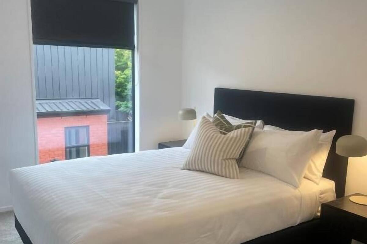 A bedroom with a large bed, a nightstand, a lamp, and a view of a building outside.