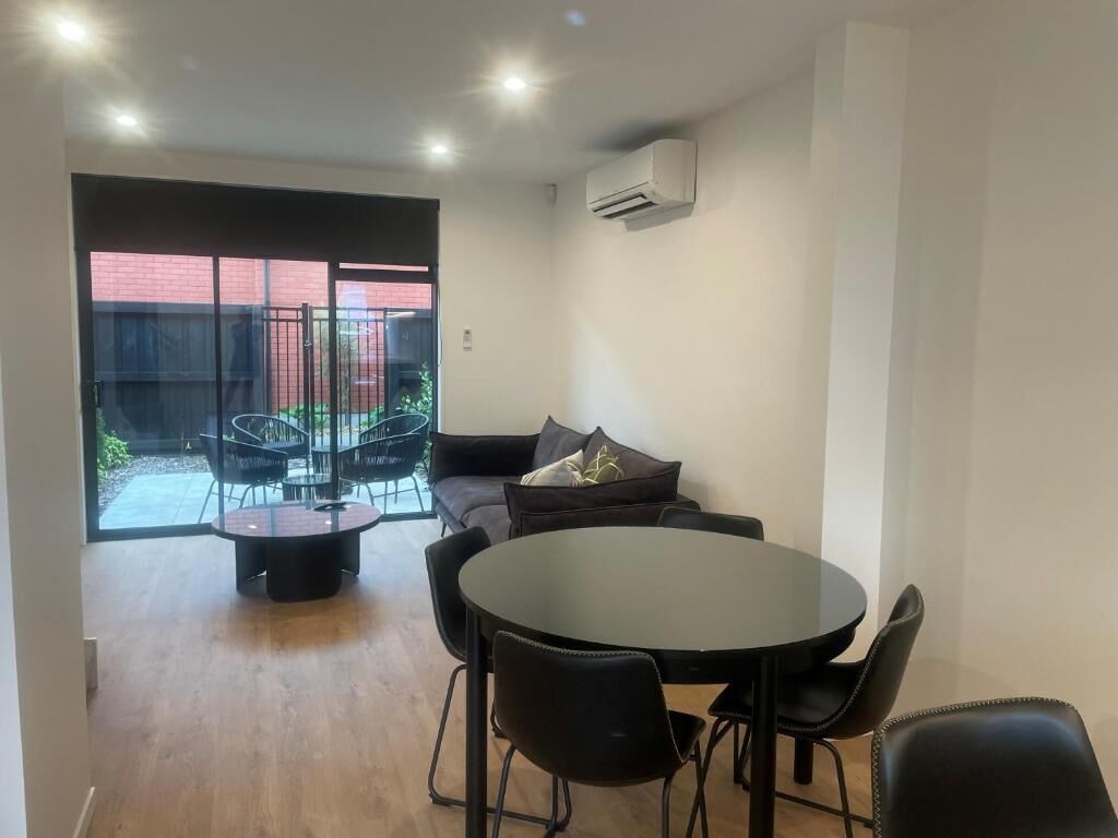 Een kamer met een ronde tafel, zwarte stoelen, een bank met kussens en een schuifdeur die uitkomt op een buitenruimte met stoelen en een tafel.