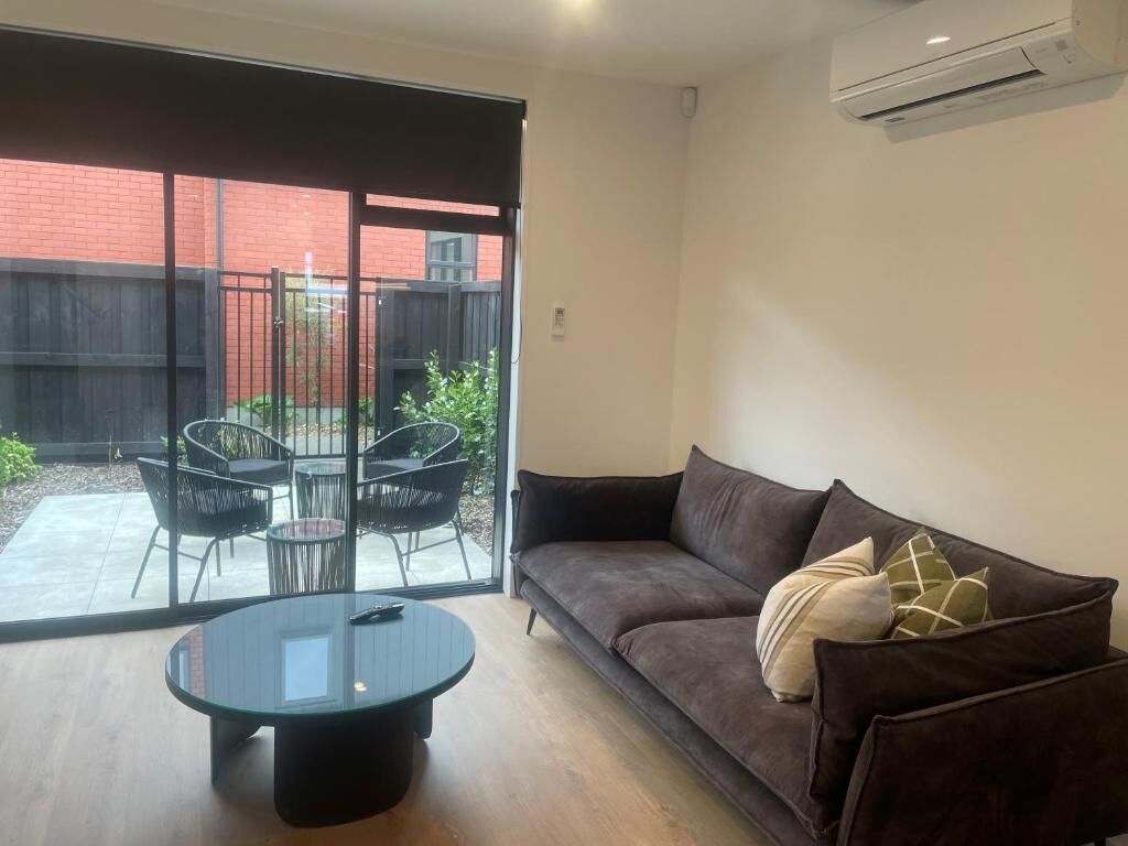 Una sala de estar moderna con un sofá, una mesa redonda y un ventanal de vidrio que da a un espacio exterior con sillas.