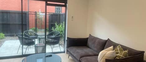 Una sala de estar moderna con un sofá, una mesa redonda y un ventanal de vidrio que da a un espacio exterior con sillas.