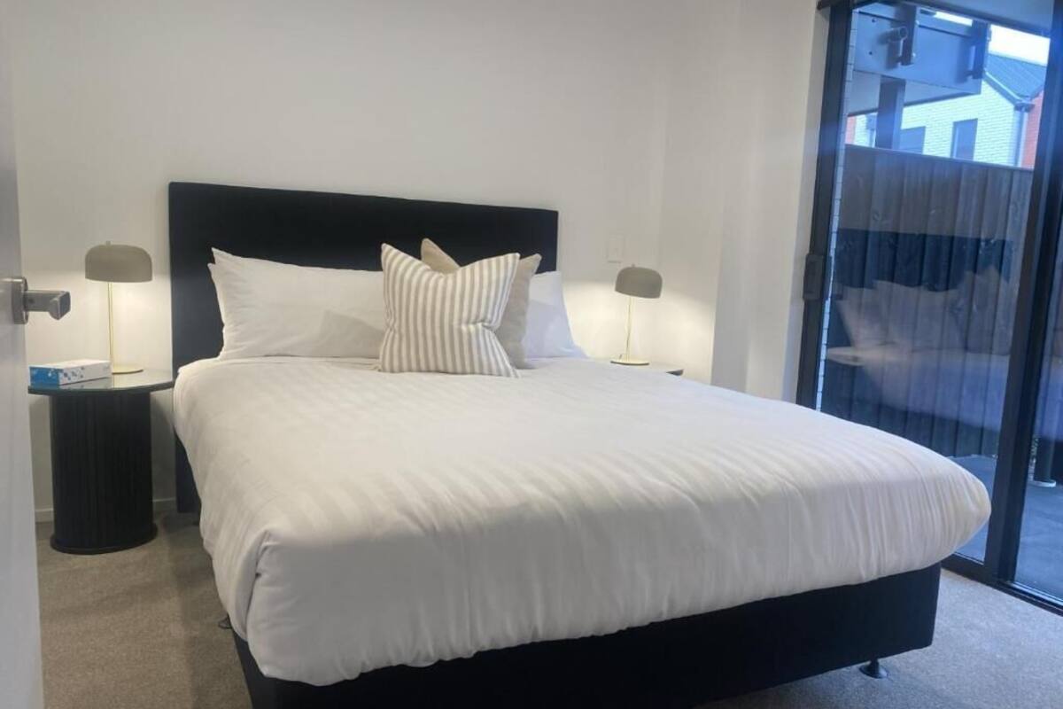 A large bed with white bedding, two bedside tables with lamps, and a view through a glass door.