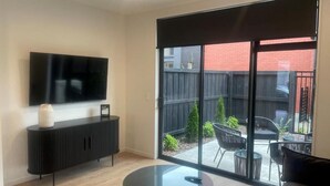 A modern living room with a flat-screen TV, a round glass table, a sofa, and a sliding glass door leading to an outdoor patio with chairs and a fence. - Merivale Executive Stays (Christchurch)