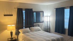 A bedroom with a bed, a nightstand, a lamp, a window with curtains, a mirror, and a patterned rug. - Spacious 4-bedroom apartment near Mayo Clinic (Rochester)