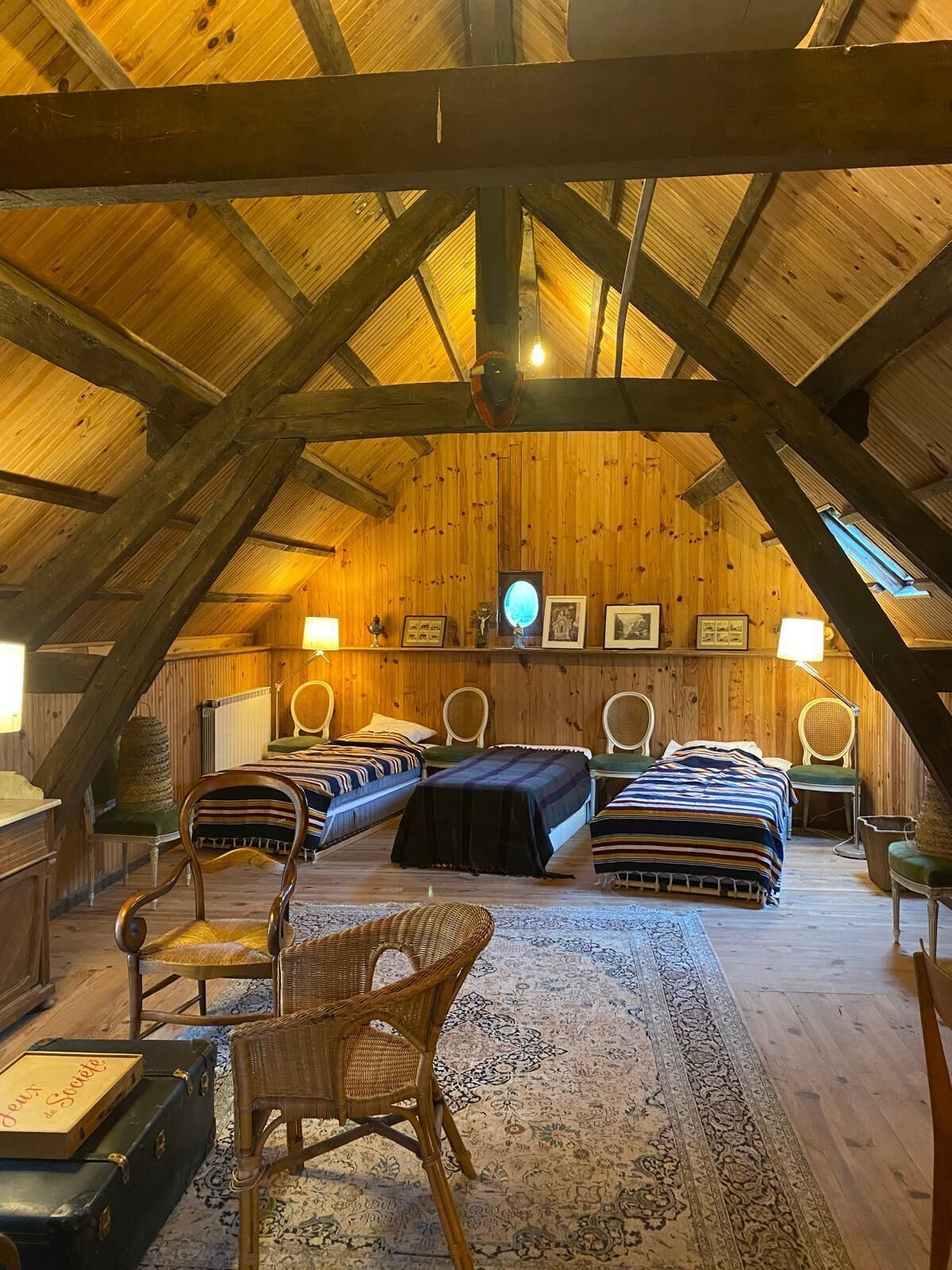 A cozy attic bedroom with wooden beams, a large bed, two smaller beds, wicker chairs, and a patterned rug.
