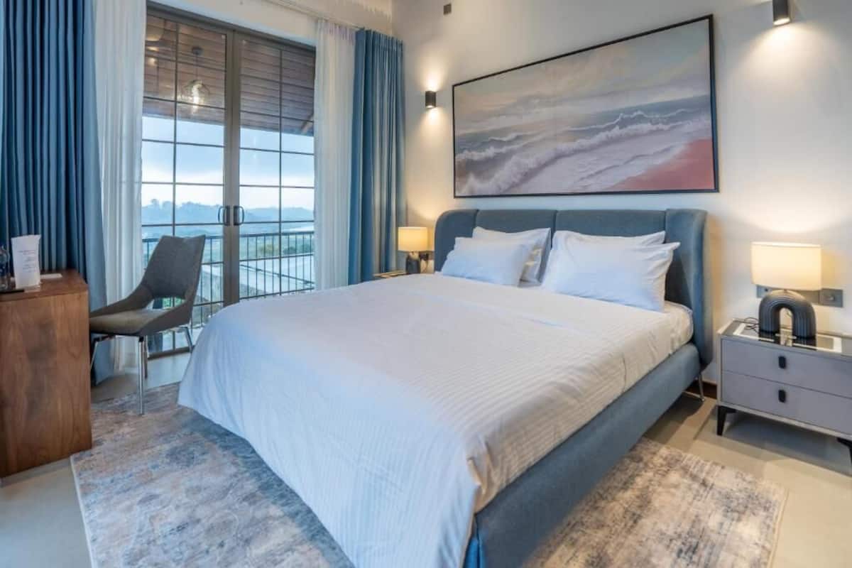 A hotel room with a large bed, a desk, a chair, a balcony with a view, and a painting on the wall.