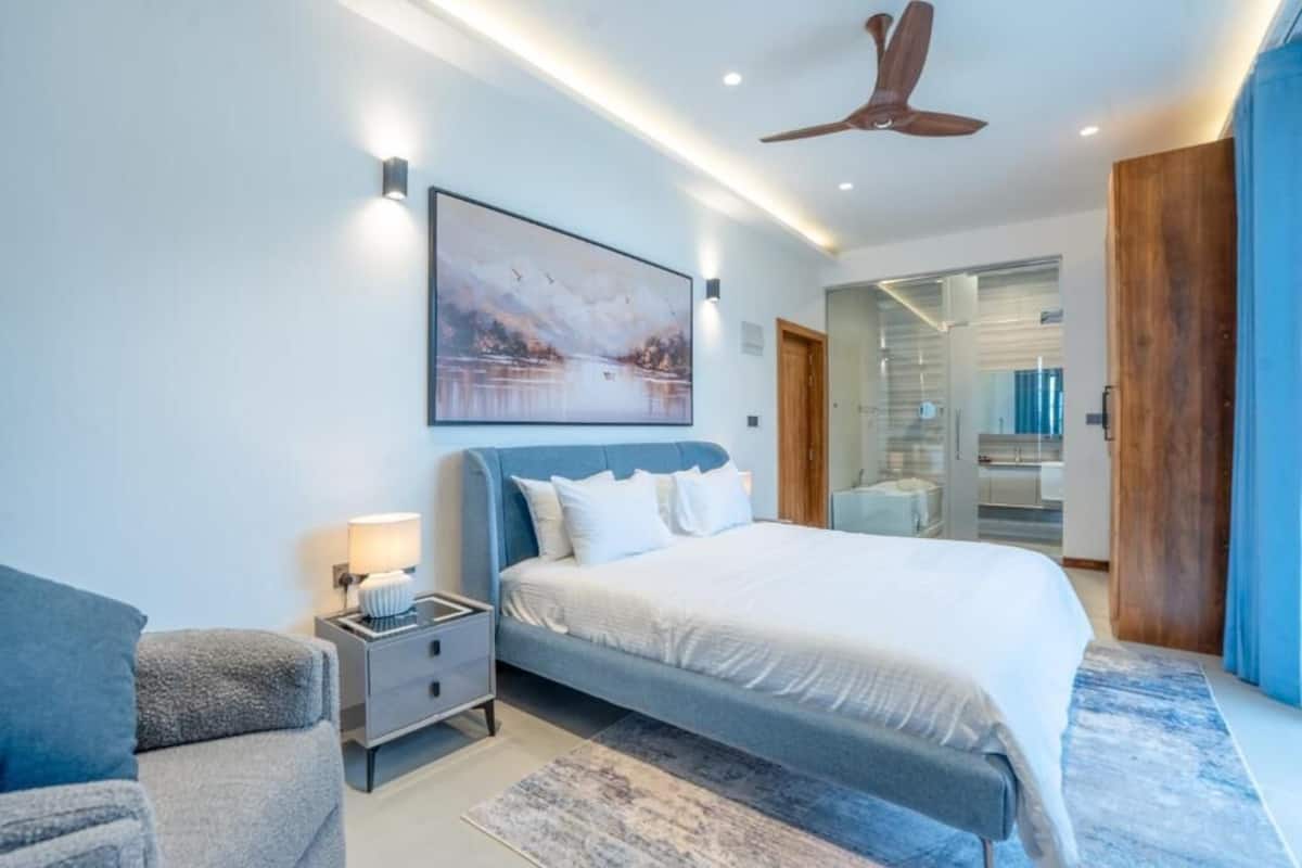 A modern bedroom with a large bed, a nightstand, a wall-mounted painting, and a ceiling fan.