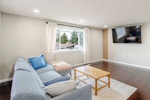 Een woonkamer met een bank, een salontafel, een aan de muur bevestigde televisie en een raam met gordijnen.