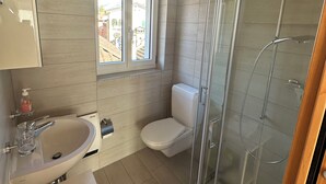 Ein Badezimmer mit Duschkabine aus Glas, Toilette, Waschbecken mit Seifenspender und Fenster mit blauem Rollo.