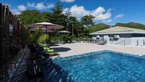 A swimming pool with lounge chairs and umbrellas, surrounded by a wooden fence and lush greenery.