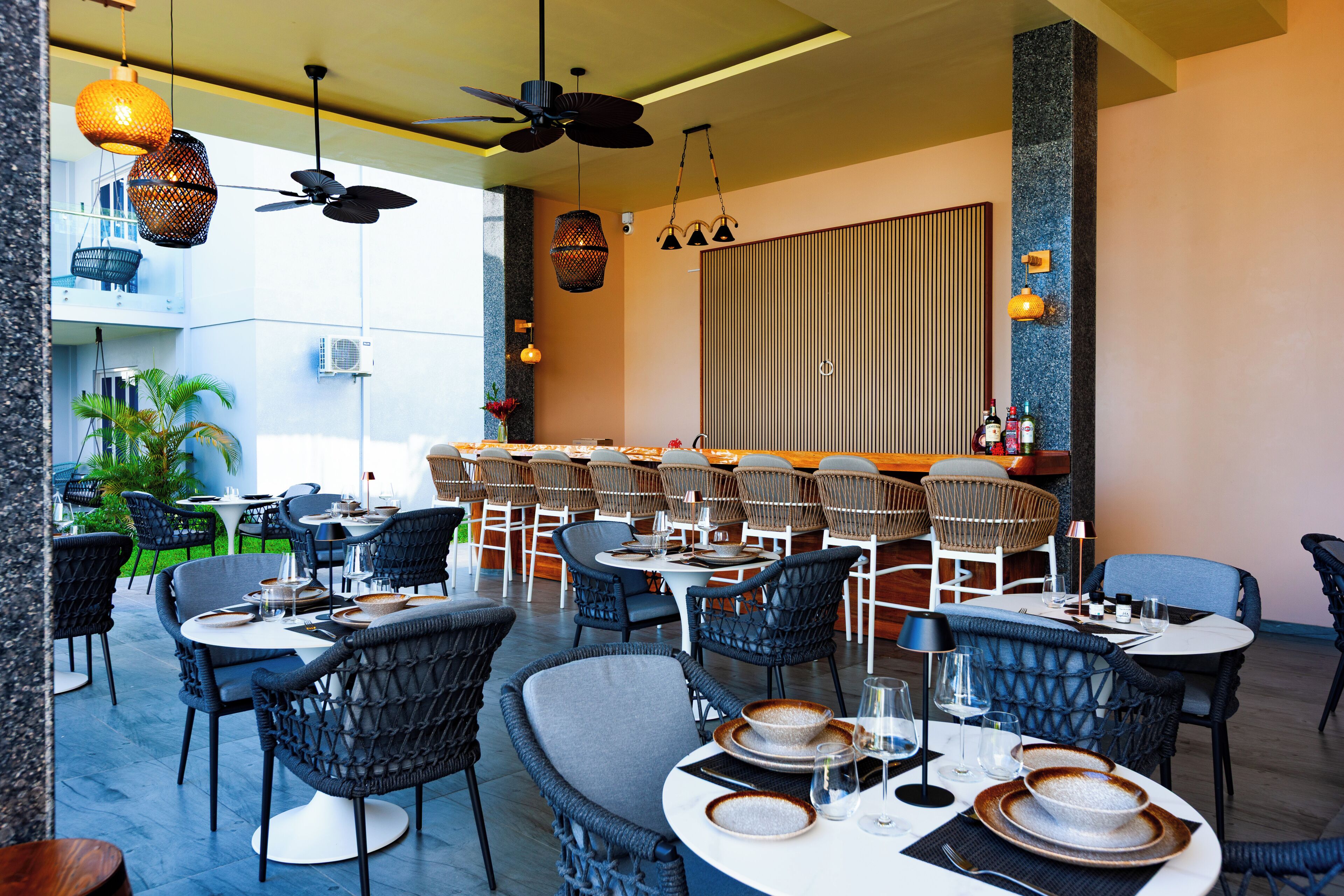 Un restaurant avec des tables rondes, des chaises en osier, des ventilateurs de plafond et des luminaires suspendus.