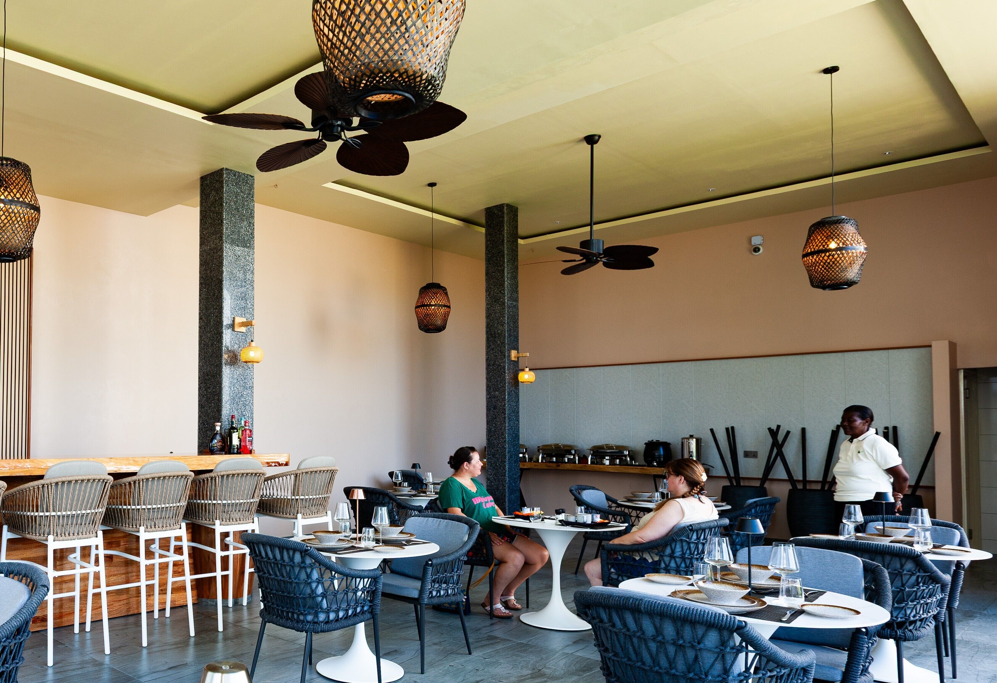 Um restaurante com cadeiras de vime, mesas redondas, ventiladores de teto e luminárias pendentes.