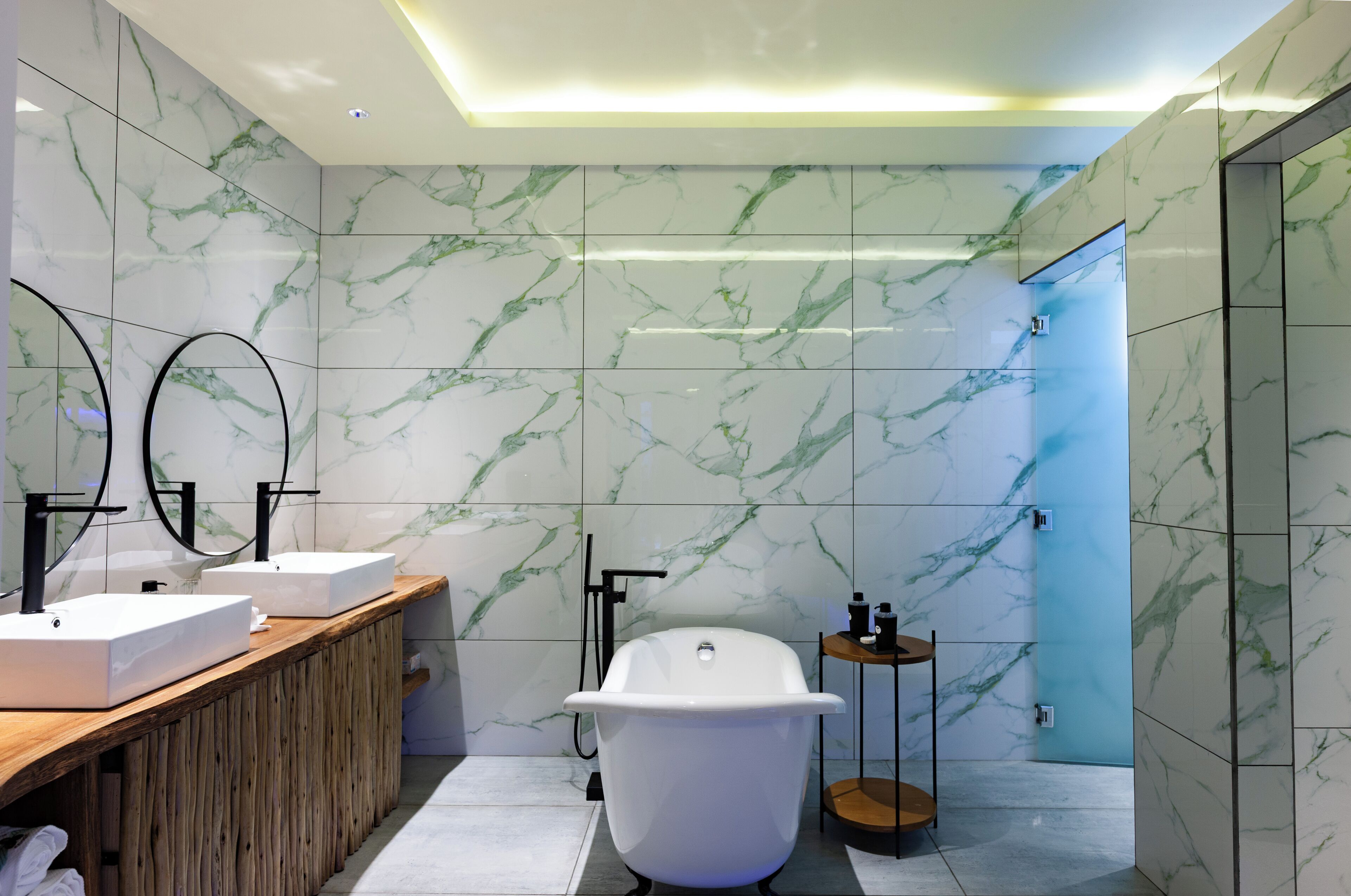 Un baño moderno con paredes de mármol, un tocador de madera, dos lavamanos, una bañera independiente y una mesita auxiliar pequeña.