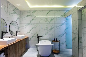 Un baño moderno con paredes de mármol, un tocador de madera, dos lavamanos, una bañera independiente y una mesita auxiliar pequeña.