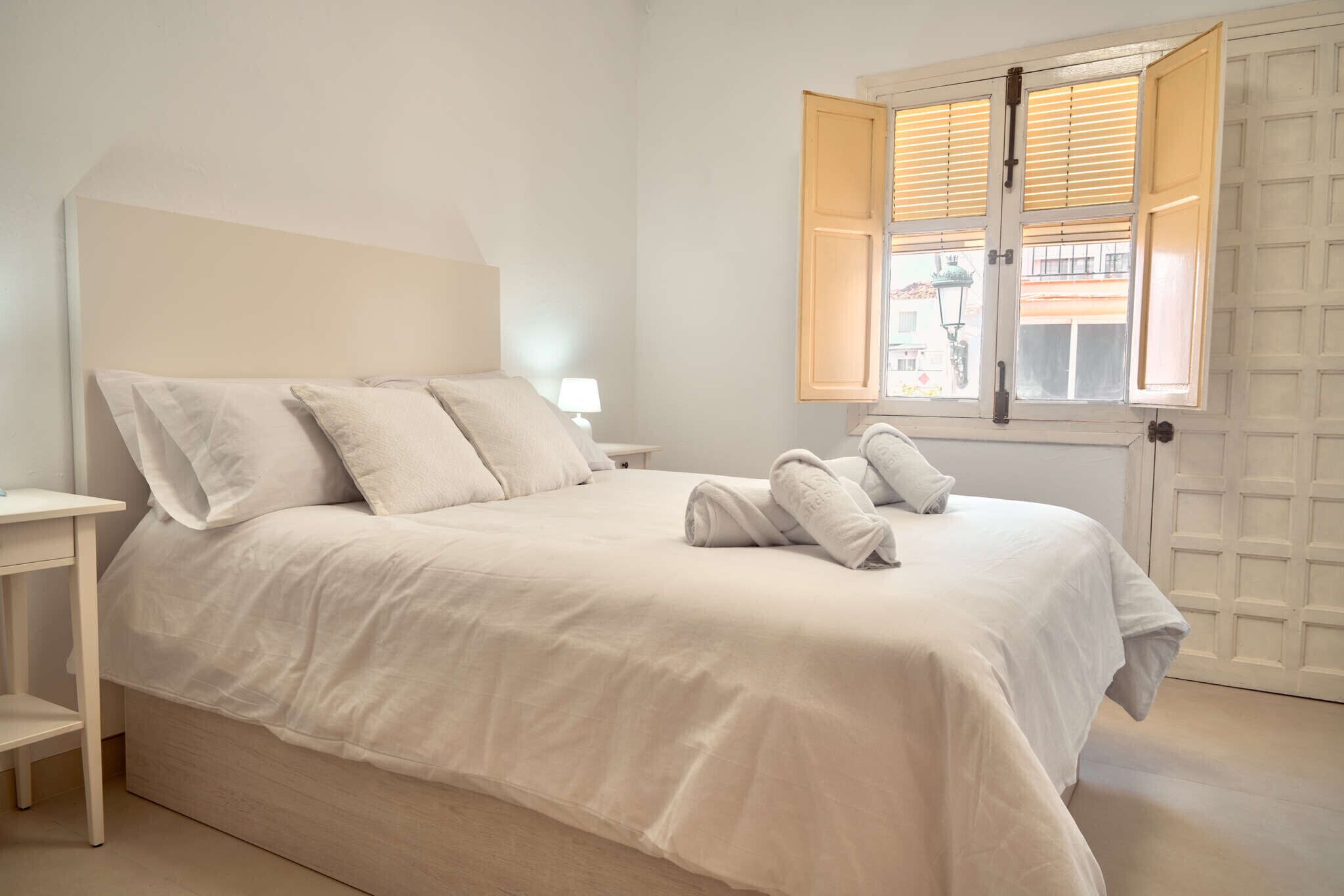 Een slaapkamer met een bed, een nachtkastje, een lamp en een raam met open luiken.