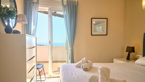 Ein Hotelzimmer mit Bett, Nachttisch, Lampe, einem gerahmten Bild an der Wand und Meerblick durch eine Schiebetür.