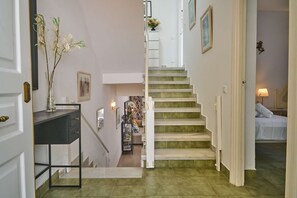 Un escalier avec une rampe blanche, un vase de fleurs posé sur une console, et un lit avec une literie blanche en arrière-plan.