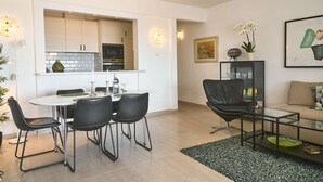 Una cocina moderna con armarios blancos, una mesa de comedor con sillas, una mesa de centro de vidrio y un sofá en una sala de estar con obras de arte y una planta.