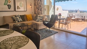 Una sala de estar con sofá, mesa de centro y sillas, un balcón con mesa y sillas, y vista a la ciudad.